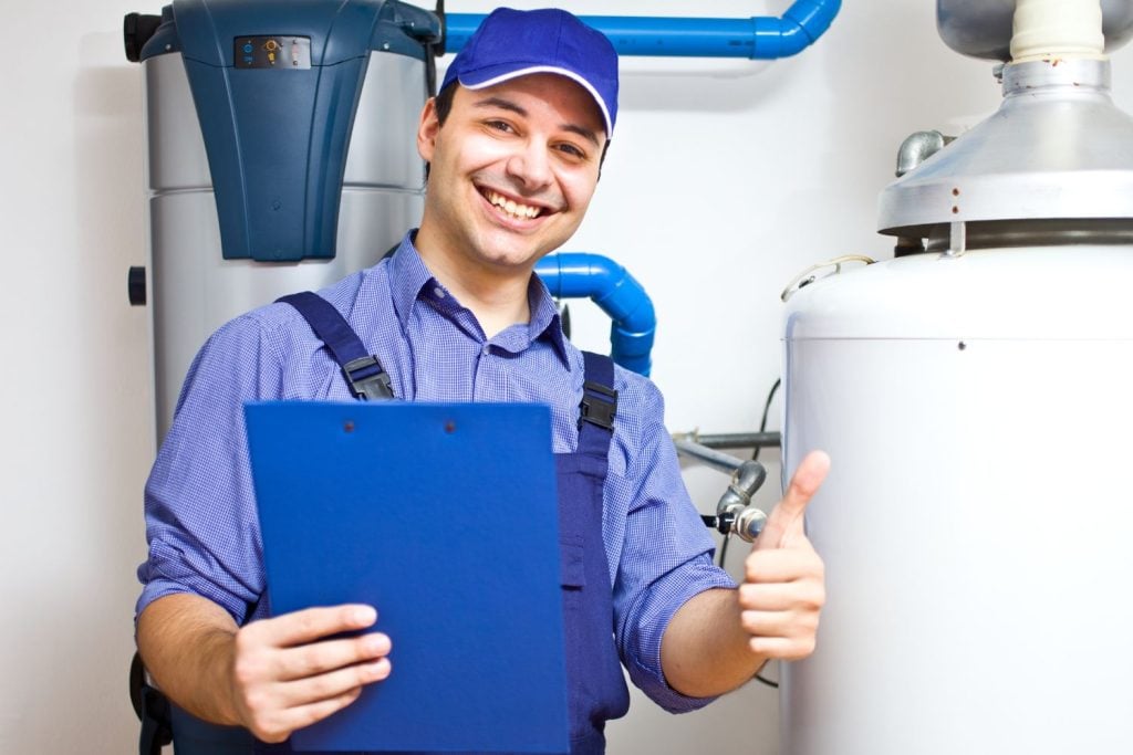 A smiling technician in overalls and a blue cap holds a blue clipboard and gives a thumbs-up next to a white water heater tank.