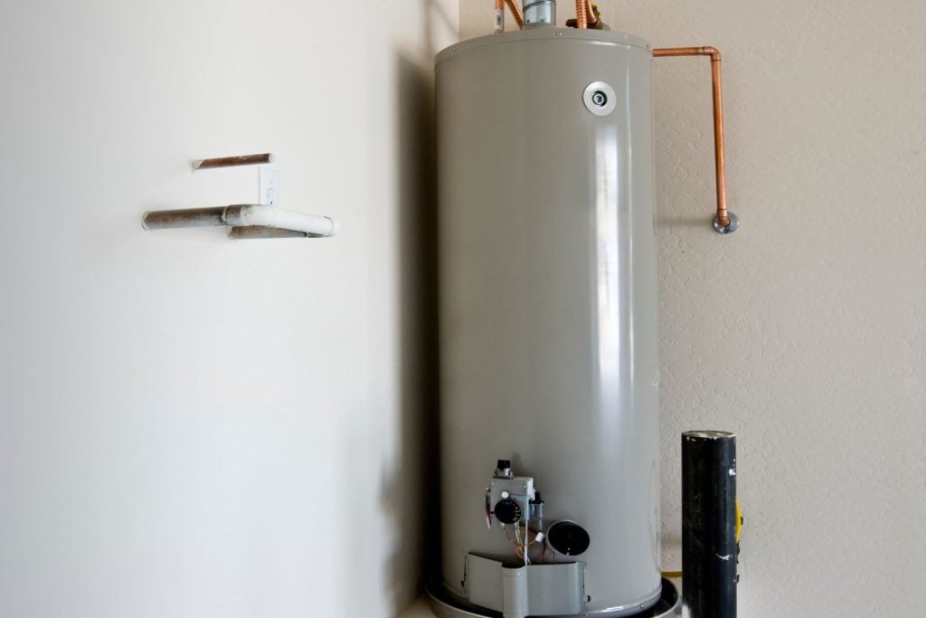 Hot Water Installation and Maintenance Carrington » Carrington A tall, cylindrical grey gas water heater stands in a corner with copper piping connected to its top and side.