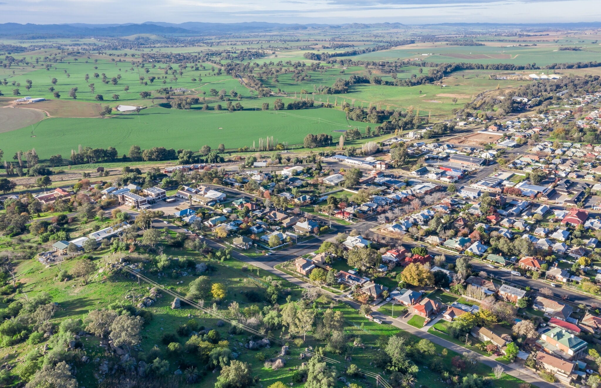 An aerial view of a rural Australian town nestled among rolling green hills and patchwork farmland