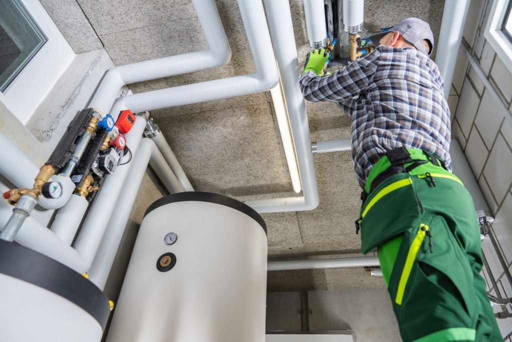Hot Water Installation and Maintenance Carrington » Carrington A technician in green work pants and a plaid shirt works on complex white insulated plumbing and valves above a large white storage tank.