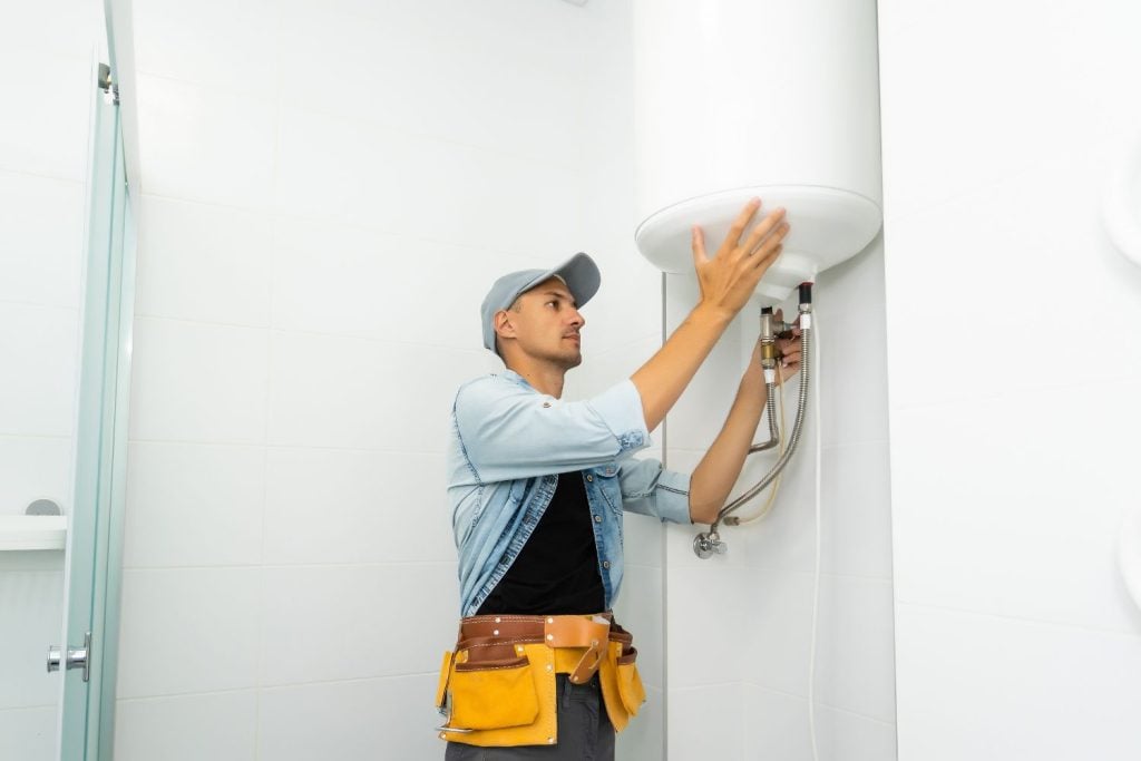 Hot Water Installation and Maintenance Bar Beach » Bar Beach A technician wearing a tool belt and a cap inspects and adjusts the bottom pipes of a large, white electric water heater.