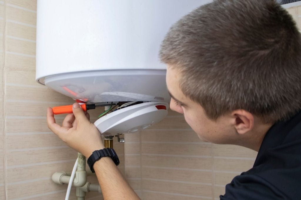 Hot Water Installation and Maintenance Beresfield » Beresfield A close-up of a young male technician with short blond hair, wearing a black shirt, using a screwdriver to perform maintenance or repair on the electrical components of a white electric water heater.
