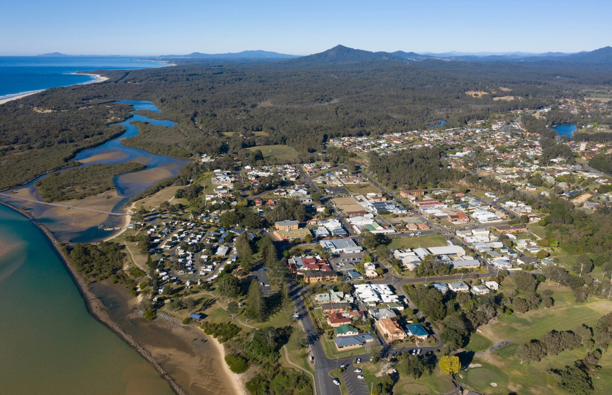 Aerial view of a coastal town surrounded by lush forest, winding waterways, and mountains in the background.