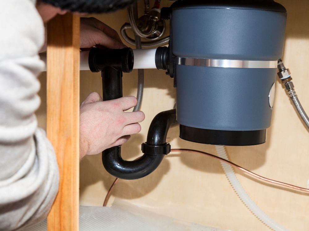 A person's hands installing a black plastic P-trap plumbing pipe connection beneath a kitchen sink.