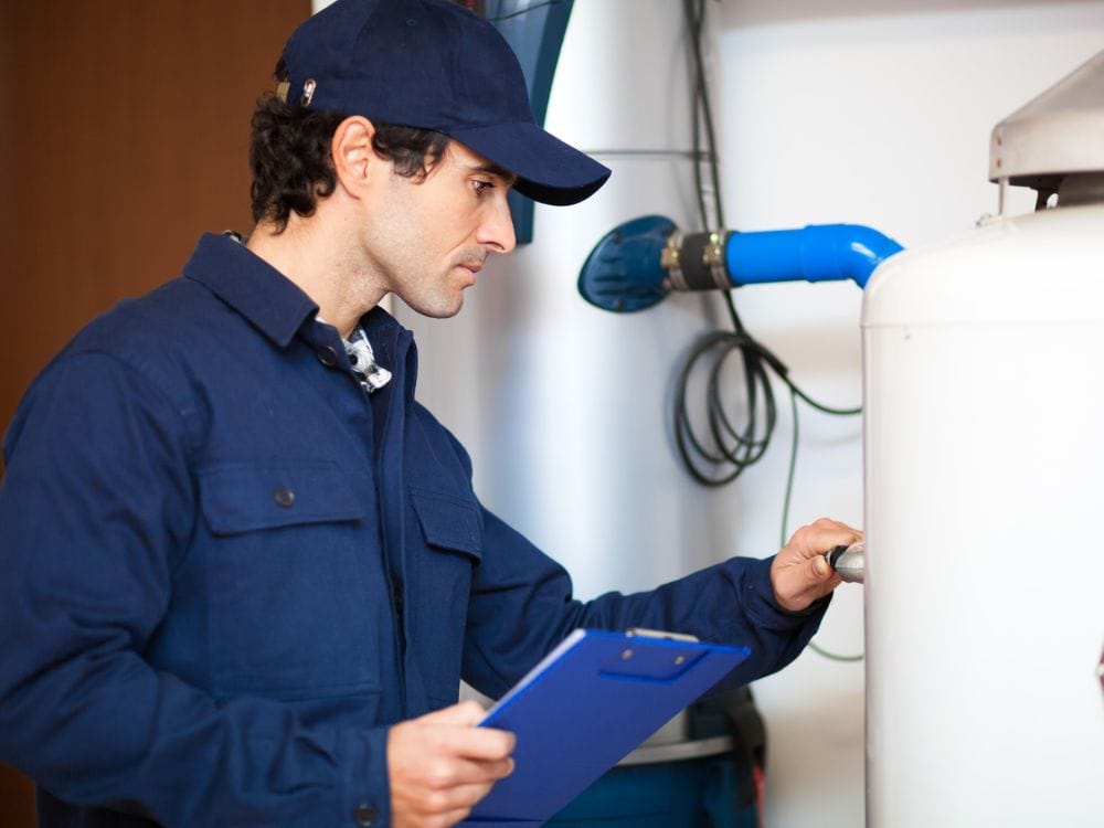 A technician wearing a blue cap and matching jacket holding a blue clipboard while inspecting a connection valve.