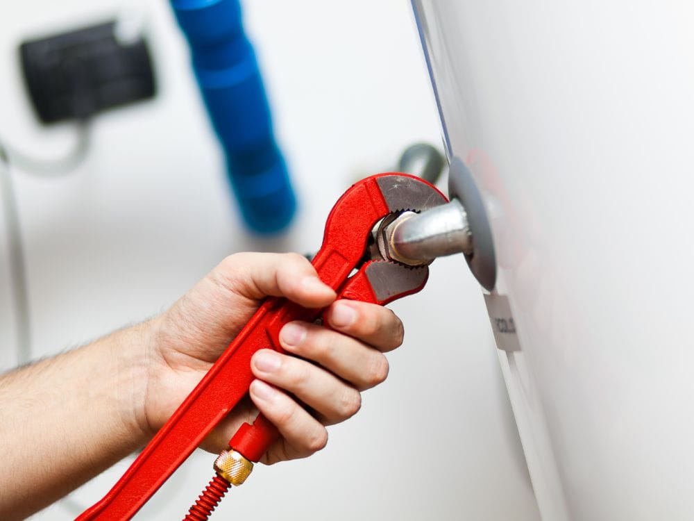 Hot Water Installation and Maintenance Lakelands » Lakelands A hand using a red plumbing wrench to tighten or loosen a metallic pipe elbow connection.