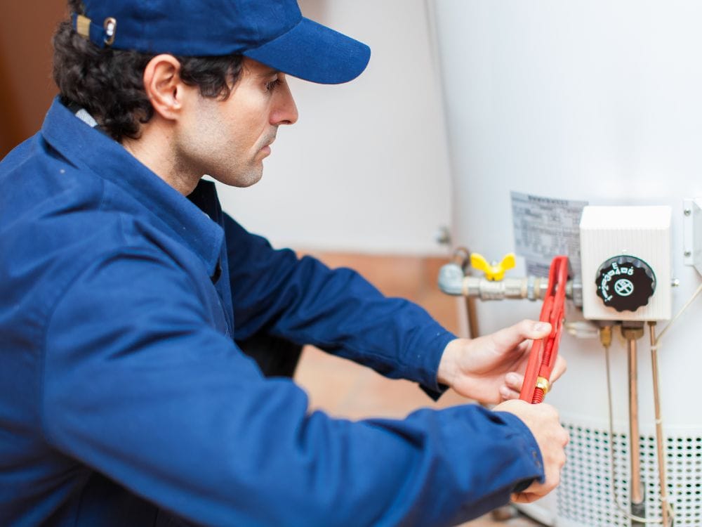 Hot Water Installation and Maintenance Lakelands » Lakelands A technician wearing a blue cap and blue shirt crouching to use a red pipe wrench on the gas valve and thermostat assembly.