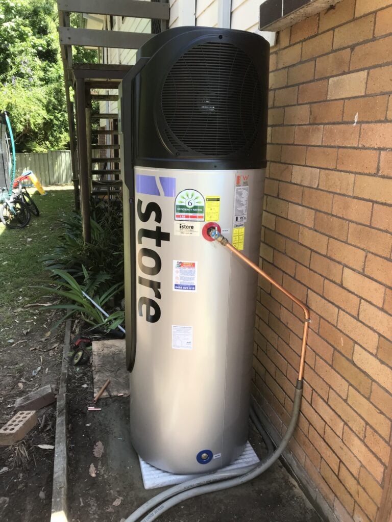 An outdoor iStore heat pump hot water system with a silver tank and black compressor unit installed against a tan brick wall.