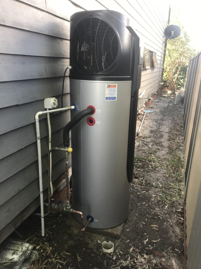 An outdoor iStore heat pump hot water system with a silver tank and black compressor unit installed on a concrete base against dark grey horizontal siding