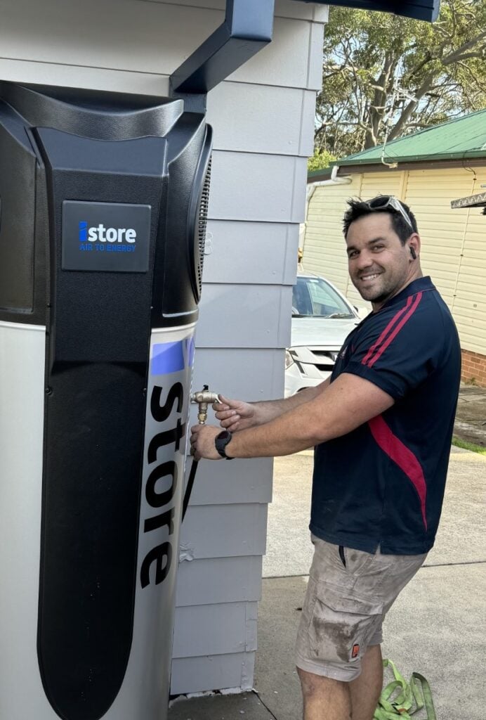 A technician smiling while installing or servicing an iStore air-to-energy heat pump system outside a residence.