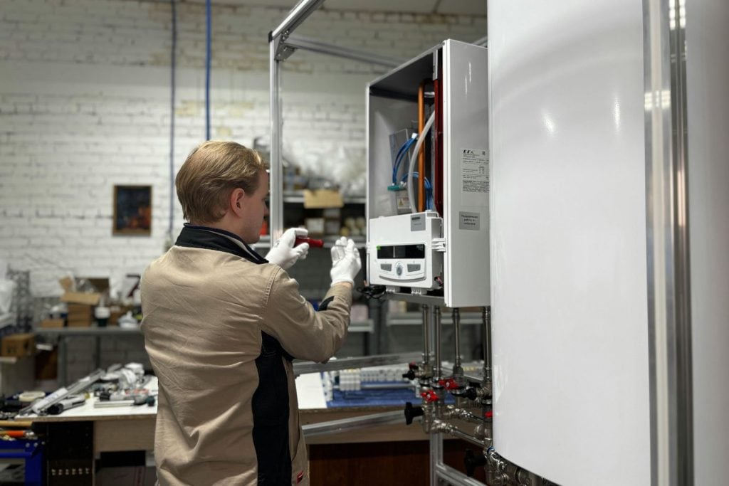 Hot Water Installation and Maintenance Jesmond » Jesmond A technician in a tan jacket and white gloves inspects the internal components of an open wall-mounted boiler in a workshop or industrial setting.