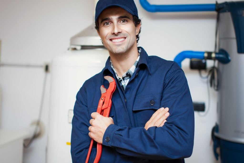 Hot Water Installation and Maintenance Jesmond » Jesmond A smiling male plumber in a blue uniform and cap, holding a red pipe wrench while standing in front of water heating equipment.