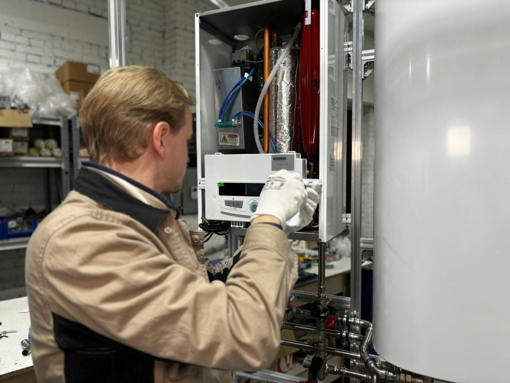 Hot Water Installation and Maintenance Mayfield » Mayfield A blond male technician in a beige work jacket and white gloves servicing the internal electronic and plumbing components of an open wall-mounted boiler system.