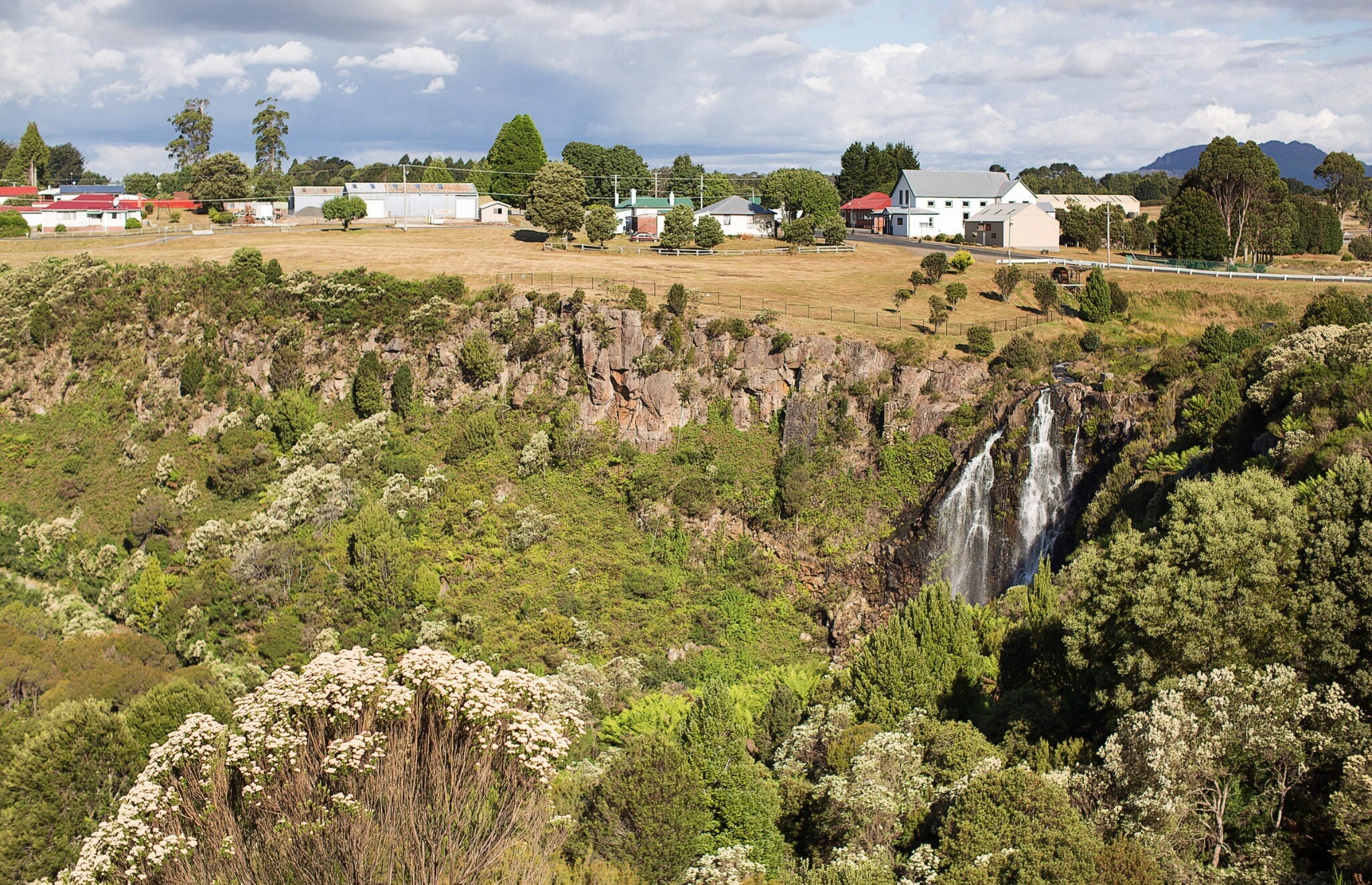 A waterfall cascading down a rocky, green cliff, with a small village and open fields visible on the plateau