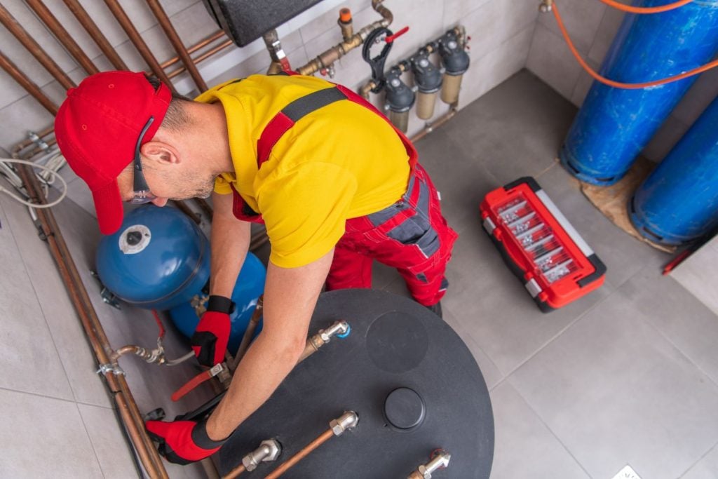A technician in a red cap and yellow shirt working on the piping of a large water heater system next to a red toolbox.