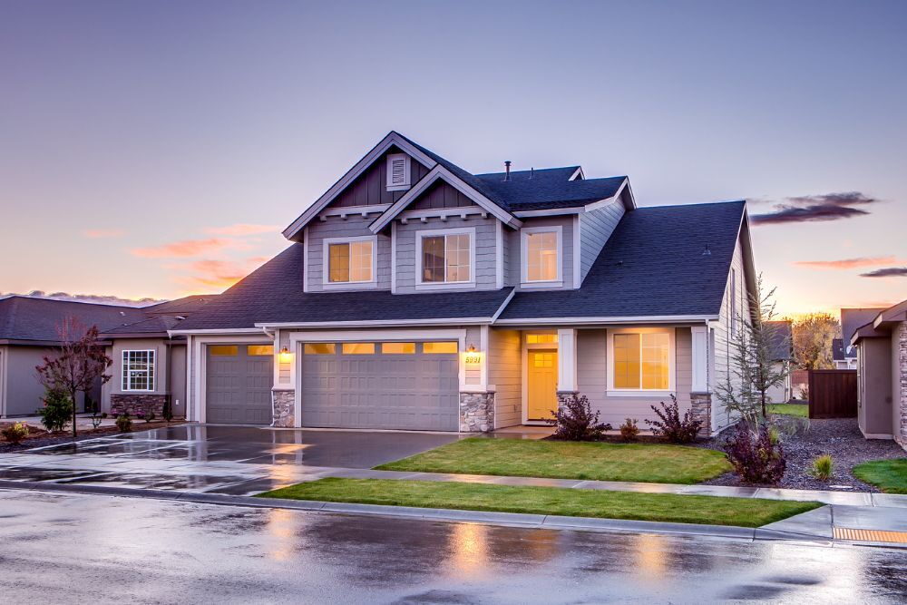 Chromagen Midea Heat Pumps » A modern two-storey suburban house with grey siding and a dark roof at sunset, featuring a wet driveway and glowing interior lights.