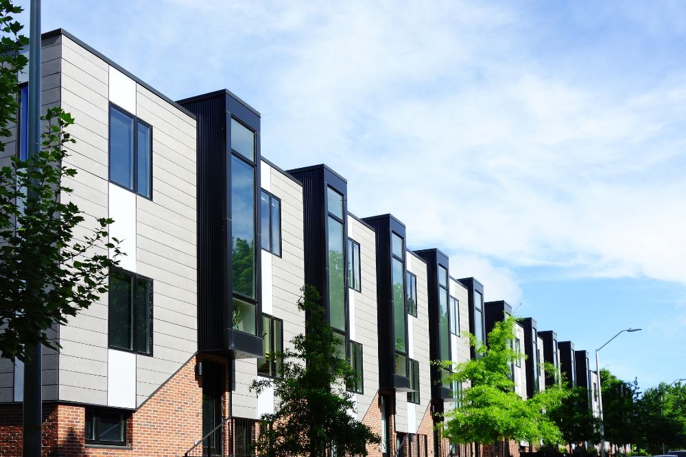 Chromagen Midea Heat Pumps » A row of modern, multi-storey townhomes with a mix of brick, light grey siding, and prominent black vertical window bays under a blue sky.