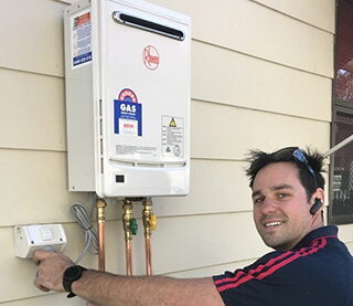 Home » Hot water system A smiling technician installs a white Rheem tankless gas water heater on an exterior wall.