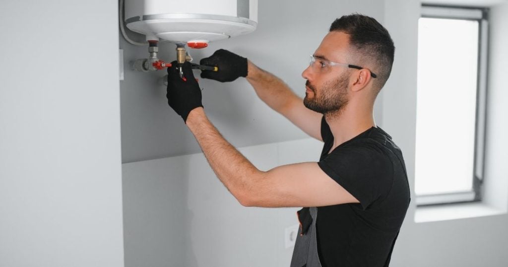 Hot Water Installation and Maintenance in Gosford » Hot Water A technician wearing safety glasses and black gloves uses a tool to adjust the water valves connected to the bottom of a wall-mounted electric water heater.