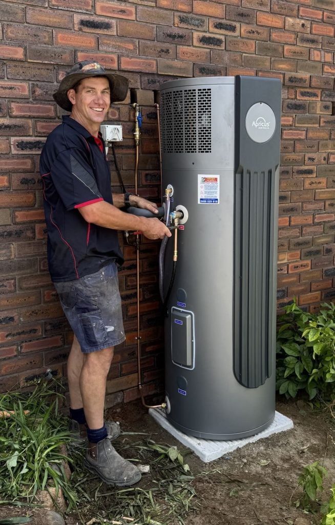 A technician in a sun hat and work clothes smiles while installing an Apricus heat pump hot water system against a brick wall.