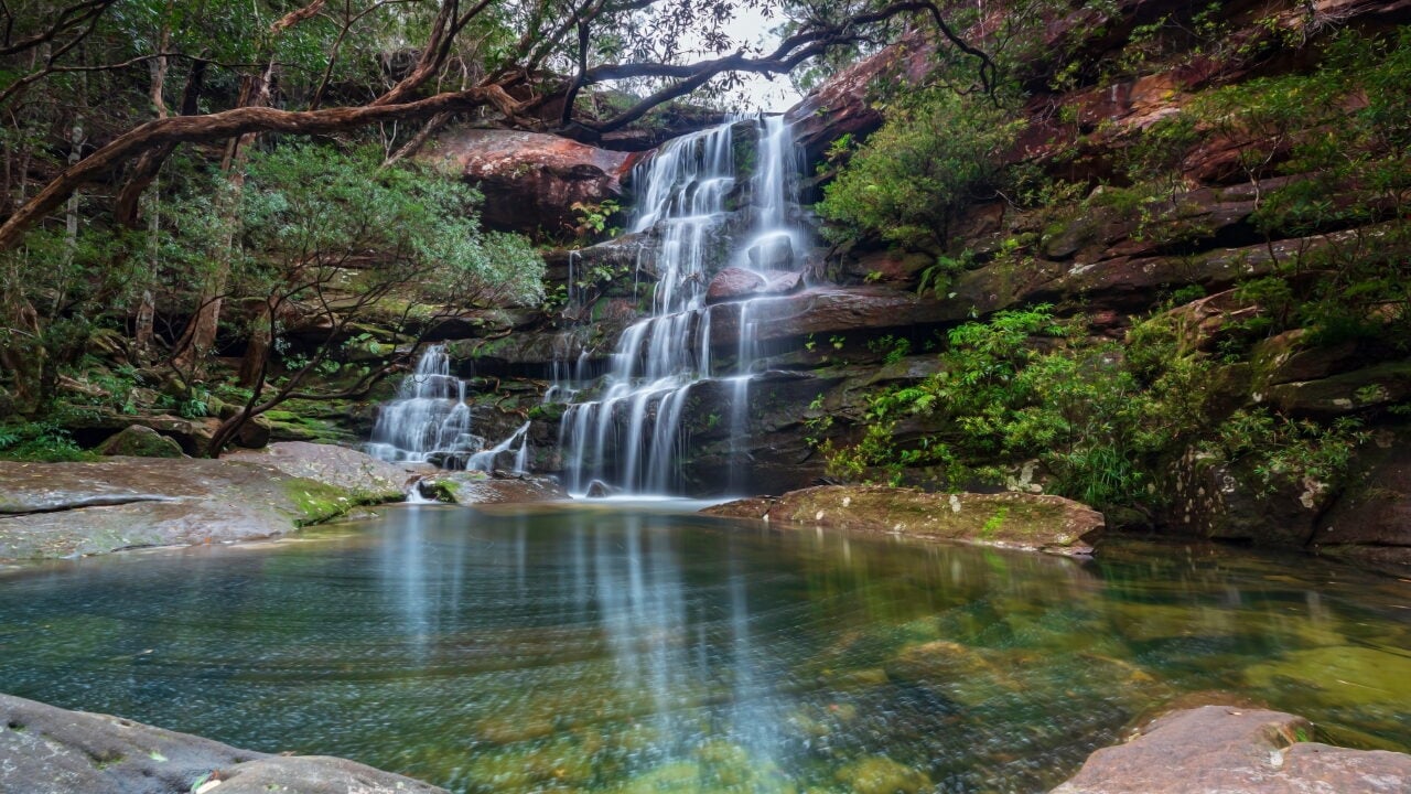 A long-exposure view of a multi-tiered waterfall cascading down a red rock face into a clear pool of Kariong