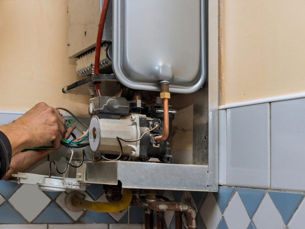 A technician's hands repairing the internal electrical wiring and circuit board panel of an opened wall-mounted gas boiler system.