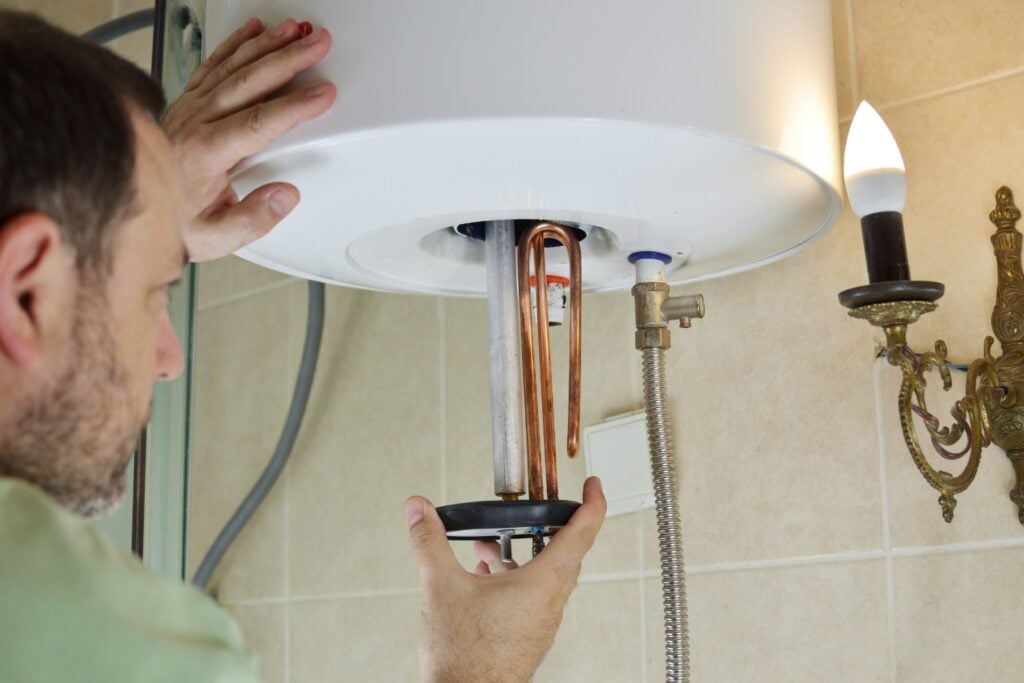 Hot Water Installation and Maintenance Phegans Bay » Hot Water A technician repairing a wall-mounted electric water heater, removing or installing a heating element assembly with copper coils from the bottom of the unit.