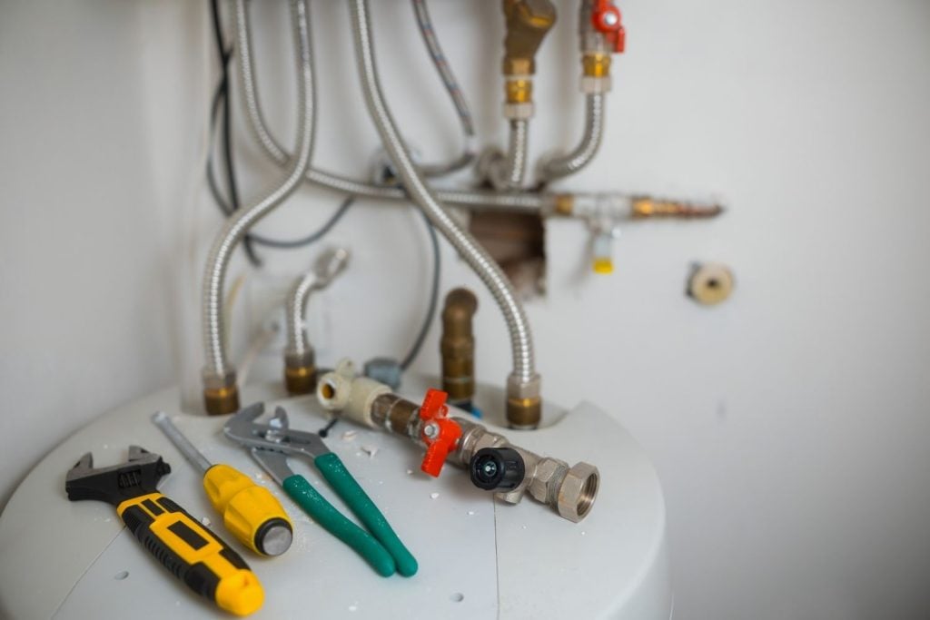 Plumbing tools, including an adjustable wrench, a screwdriver, and pliers, lying on top of a water heater with flexible metal pipes in the background.