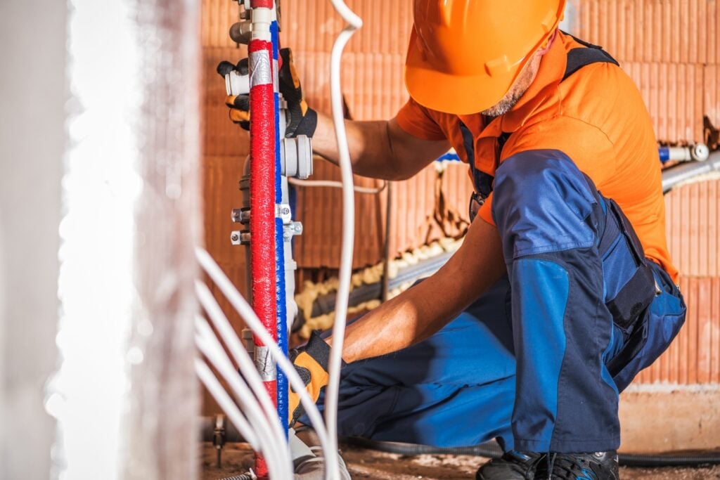 Hot Water Installation and Maintenance Phegans Bay » Hot Water A plumber wearing an orange hard hat, an orange shirt, and blue overalls, crouching down to install or repair insulated water pipes inside a building under construction.