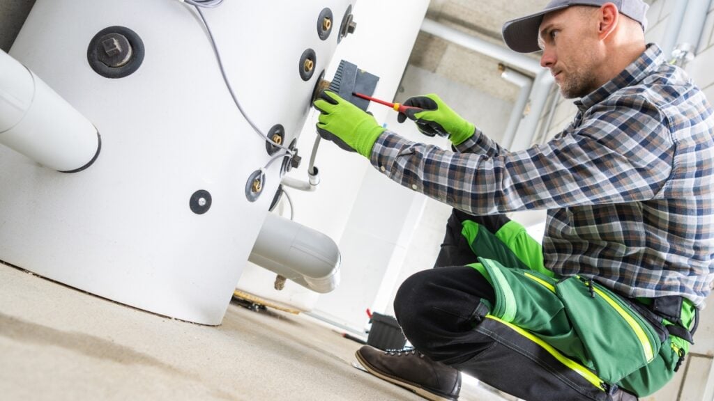 A technician in a plaid shirt and green work trousers kneels to inspect and adjust a large white hot water system