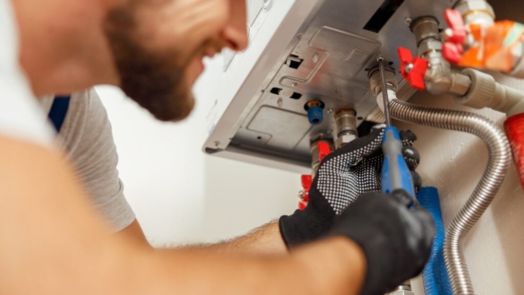 A technician wearing black gloves uses a screwdriver to perform maintenance on a wall-mounted hot water system