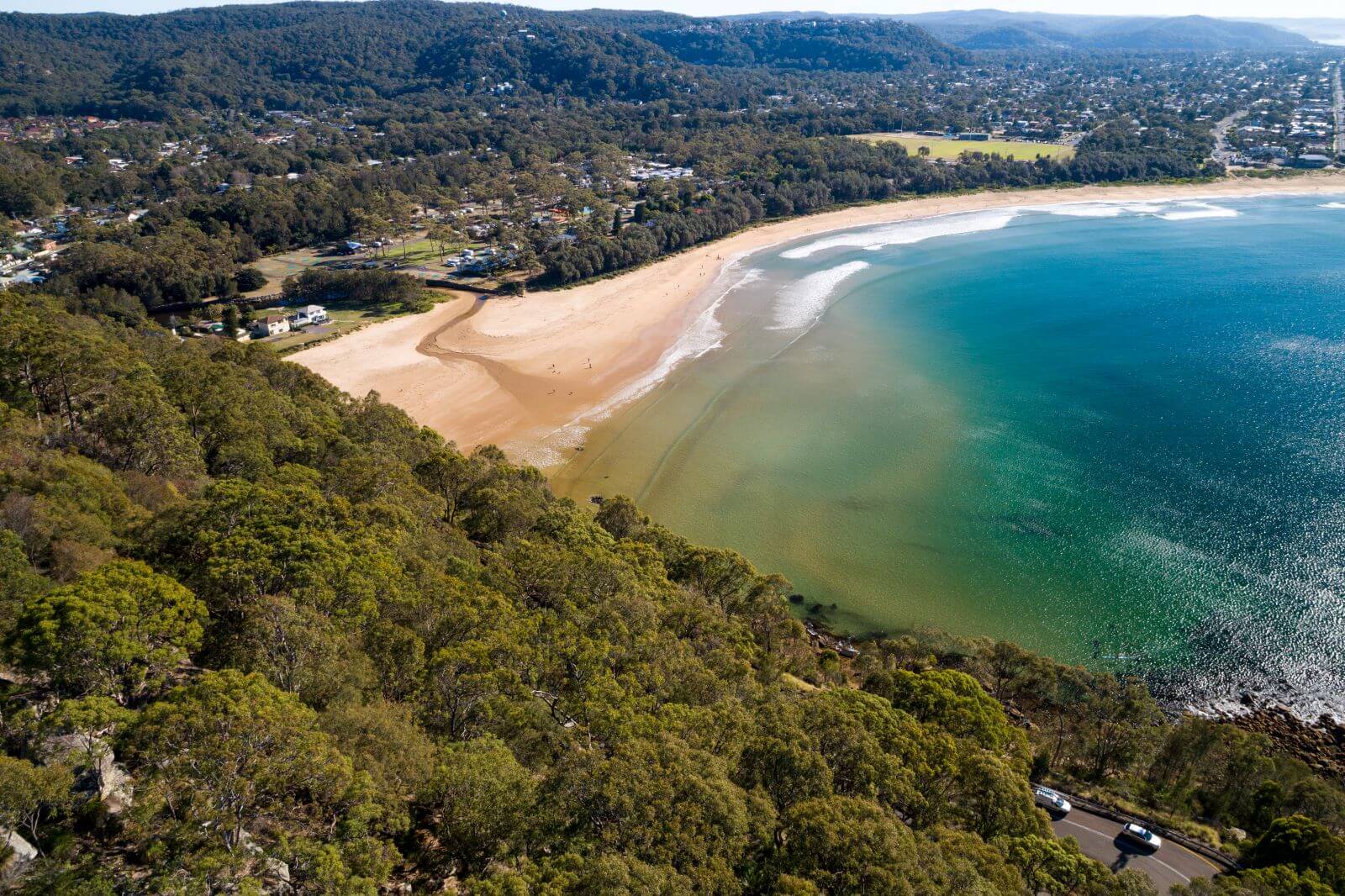 An aerial view looking down from a heavily forested hill toward a sweeping sandy Umina beach.