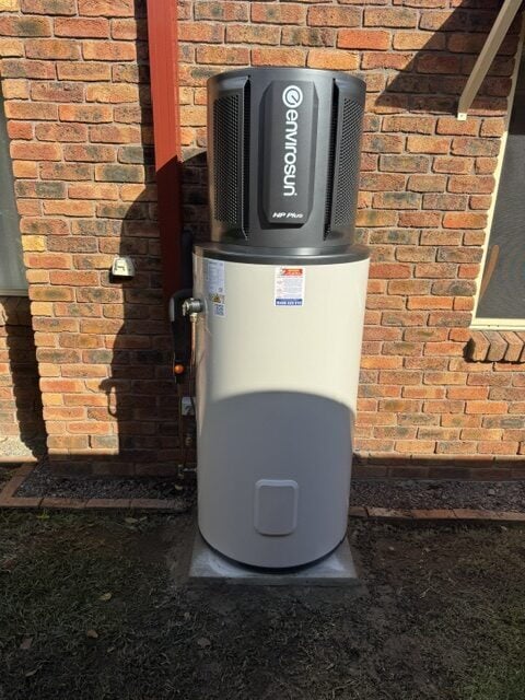 Envirosun HP Plus heat pump water heater installed outdoors on a concrete pad against a brick wall.