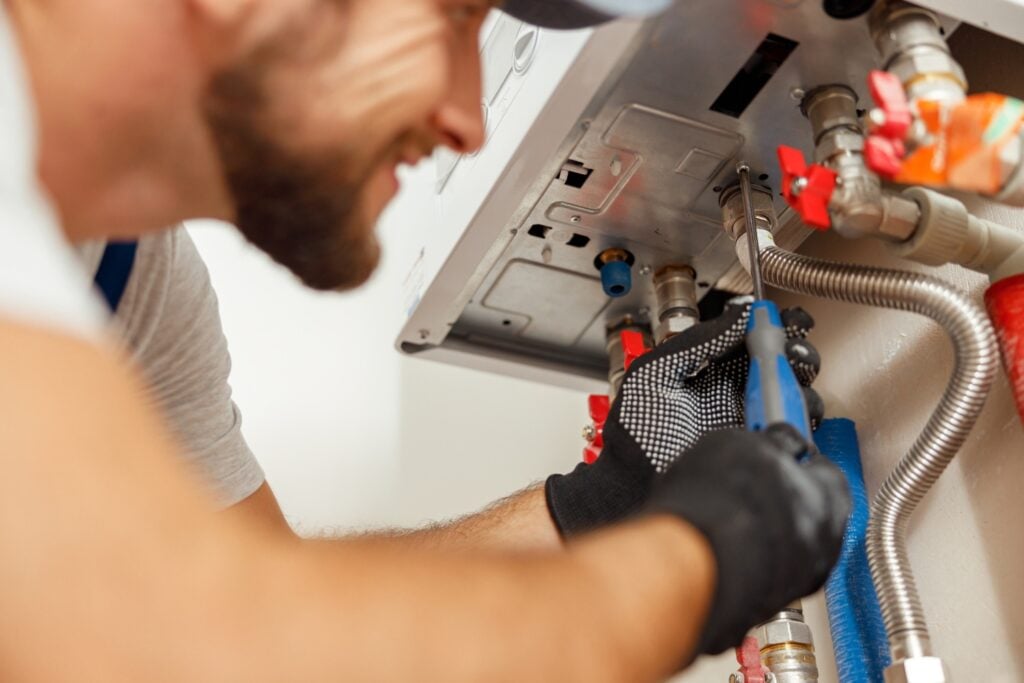 A technician wearing black gloves and using a screwdriver to repair or install a wall-mounted water heater system.