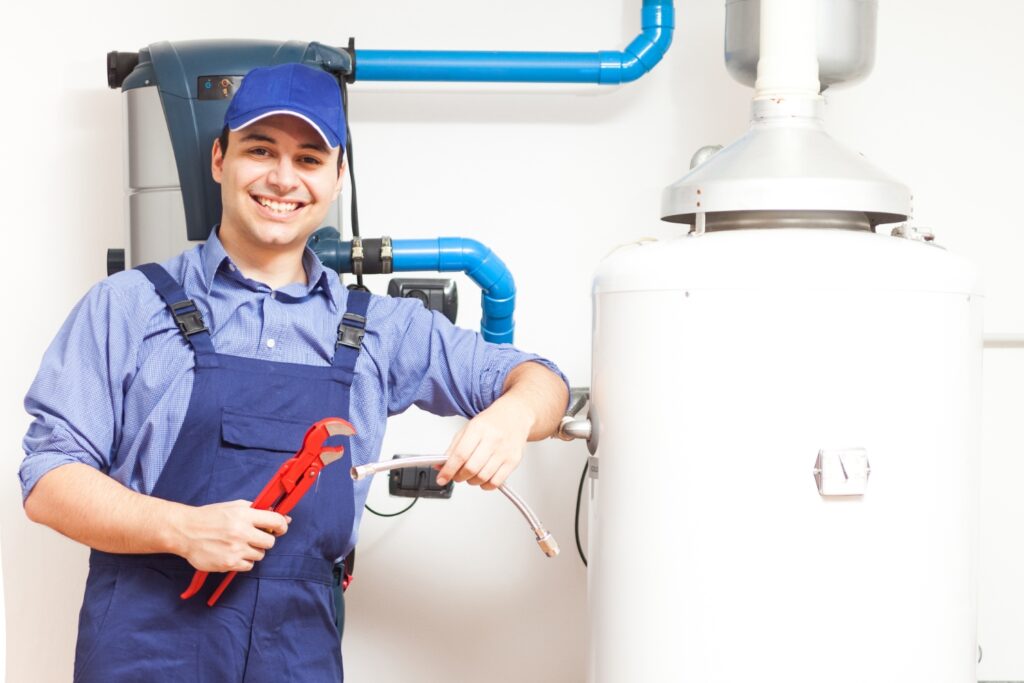A smiling plumber in blue overalls and a cap holding a pipe wrench and a flexible hose next to a large water heater tank.