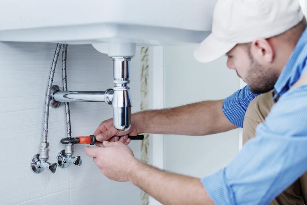 A plumber using a wrench to tighten or adjust the pipe fittings under a white bathroom sink.