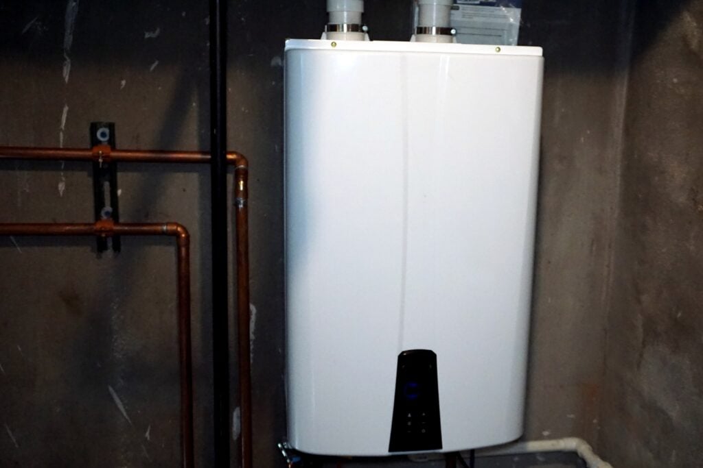 A modern white tankless water heater or heat pump control unit mounted on a concrete wall next to copper plumbing pipes.