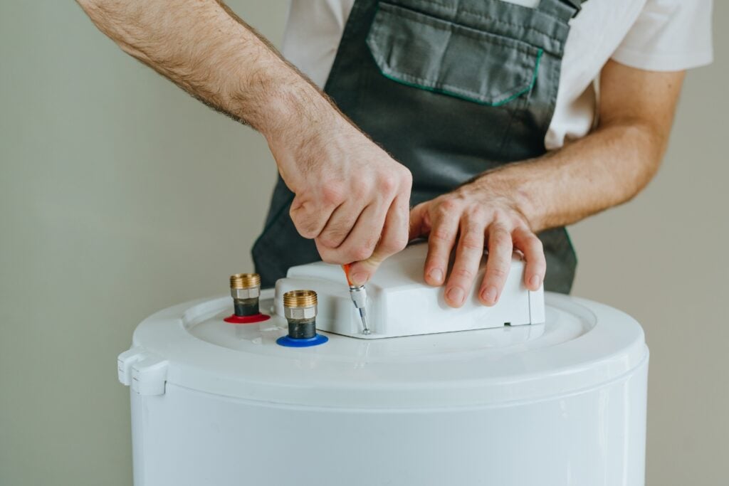 A technician in grey overalls using a screwdriver to secure a white component on top of a cylindrical water heater tank.