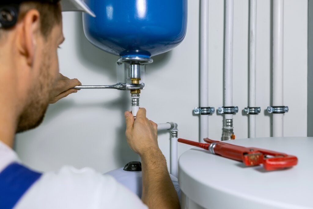 A plumber using a wrench to install or repair a pipe fitting connected to a blue expansion tank on a water heating system.
