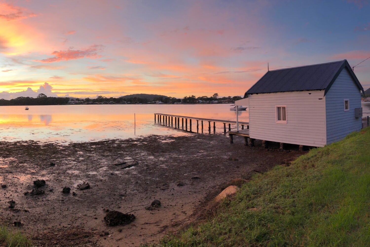 A scenic sunset view over a calm bay during low tide, featuring a small white boatshed on stilts with a wooden jetty extending into the reflective orange and pink water.