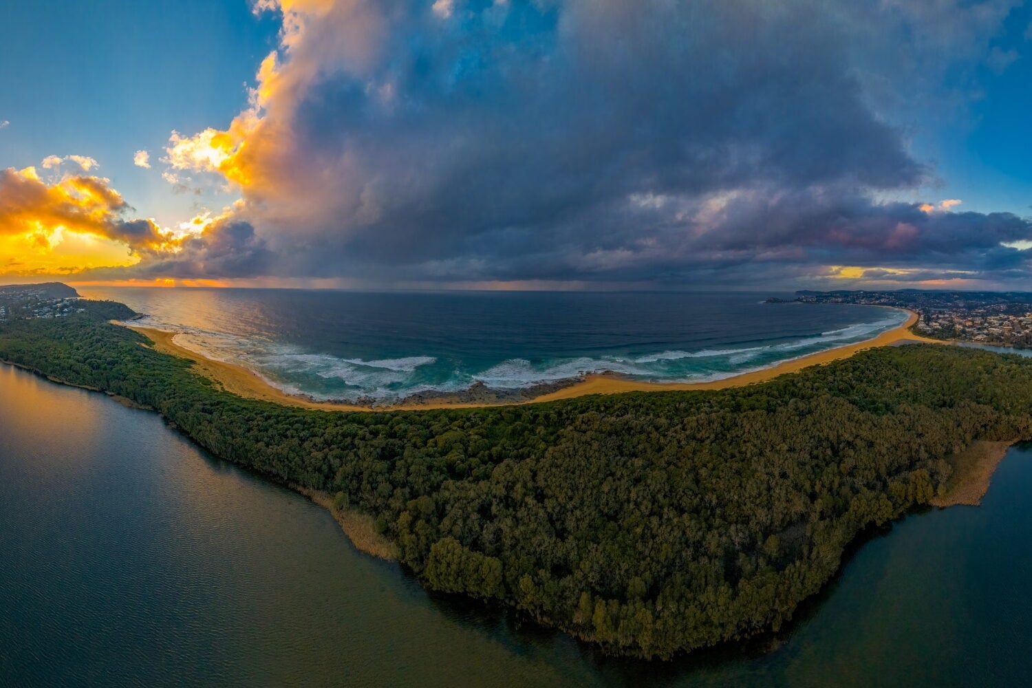 A crescent-shaped sandy beach and ocean bay separated from an inland waterway by a thick forest, under a dramatic sunrise sky with dark storm clouds.