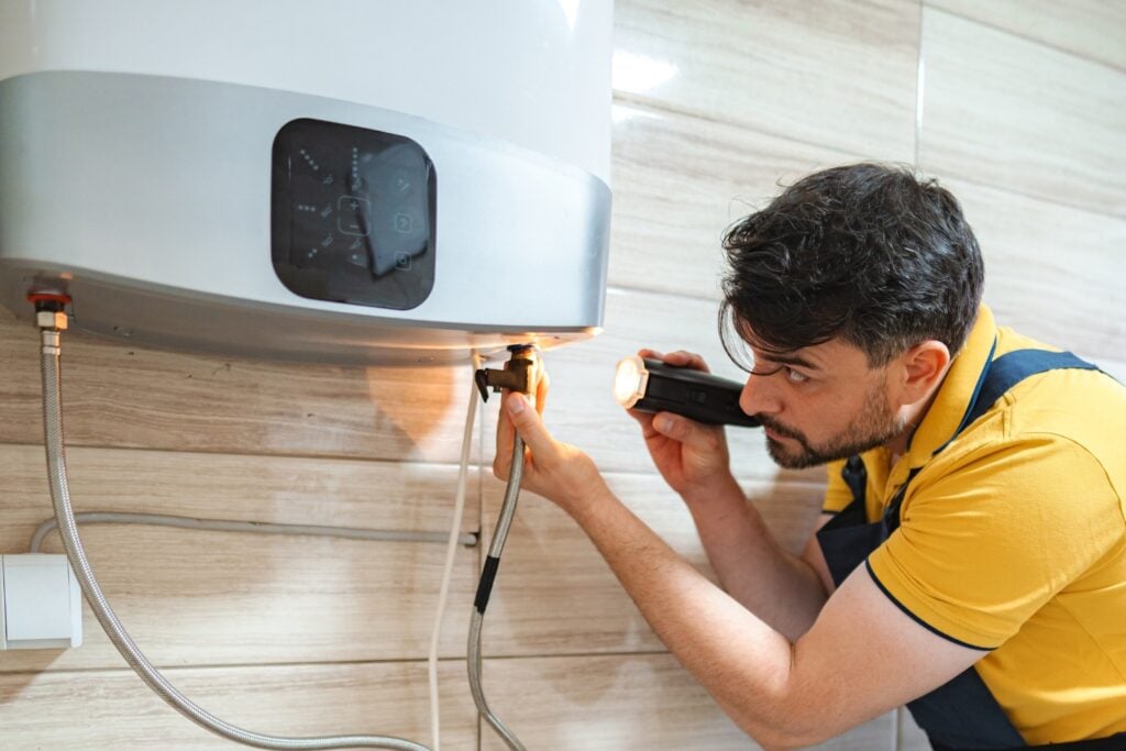A technician in a yellow polo shirt shining a flashlight onto the connection valves underneath a modern tankless water heater.