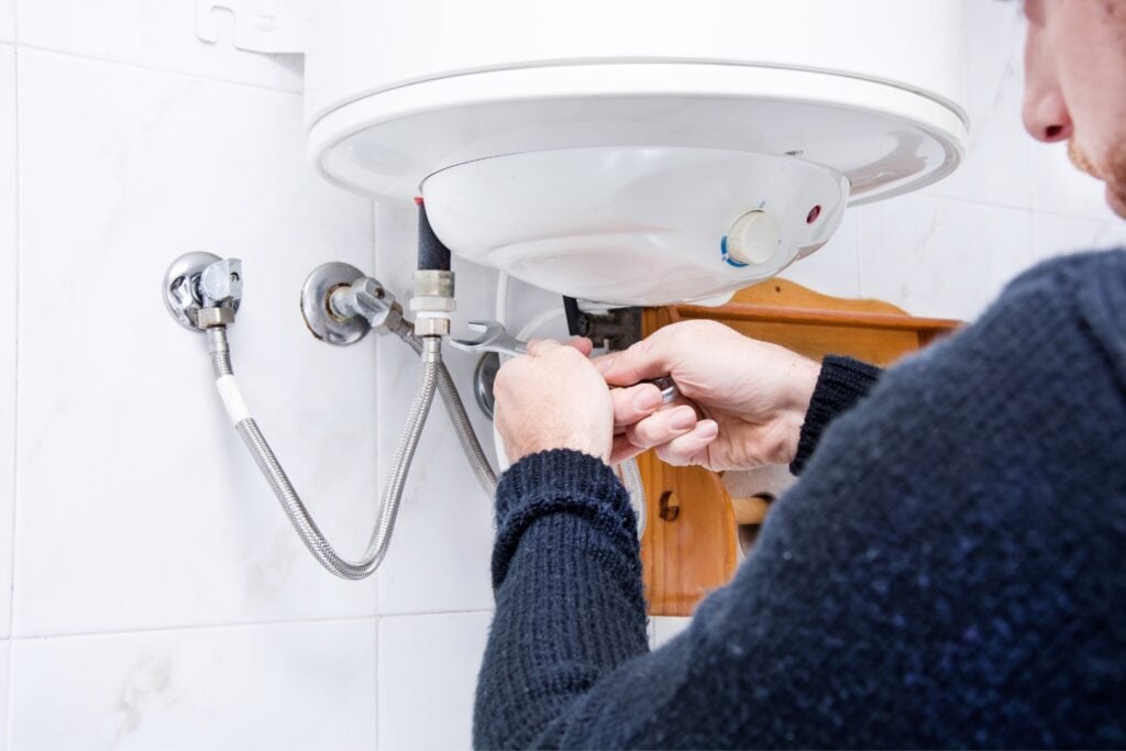 A technician using a small spanner wrench to adjust or repair the pipe connections underneath a white water heater unit mounted on a tiled wall.