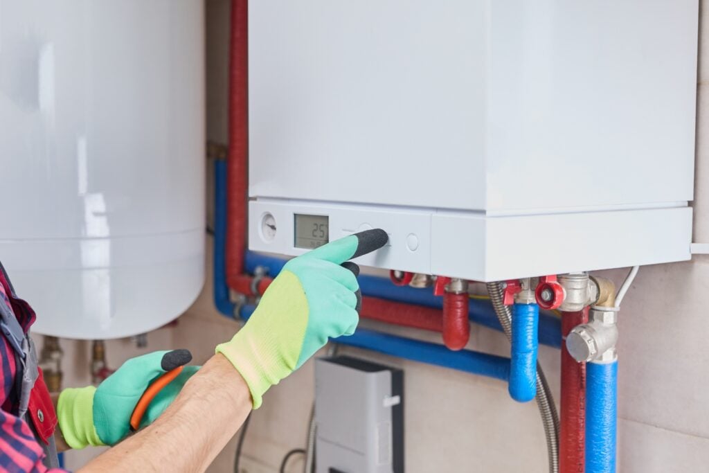 A close-up of a technician wearing green and yellow work gloves adjusting a button on a modern wall-mounted boiler system next to exposed red and blue piping.