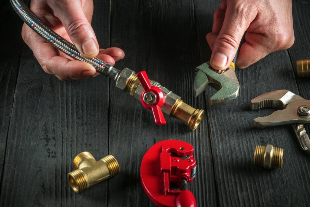 A close-up of hands connecting a flexible braided hose to a brass valve, surrounded by plumbing tools like an adjustable wrench and pipe cutter on a dark wooden surface.