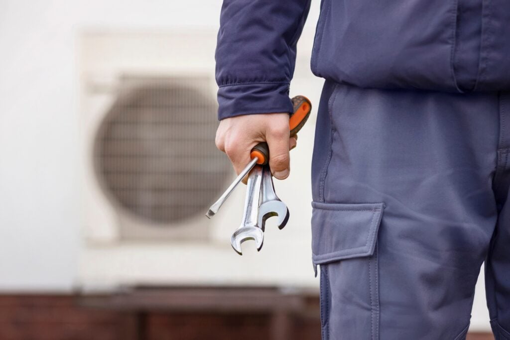 A technician in blue workwear holding a screwdriver and two metal wrenches.