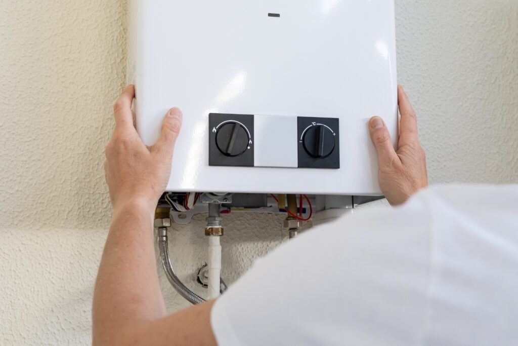 A close-up view from behind a technician holding or installing a white wall-mounted water heater with two black control dials on the front panel.