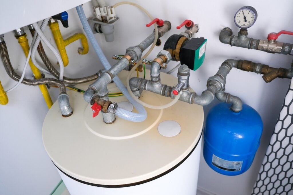 A high-angle, close-up view of the top of a white water heater tank showing connected metal pipes, valves, a green pump component, and a blue expansion tank.