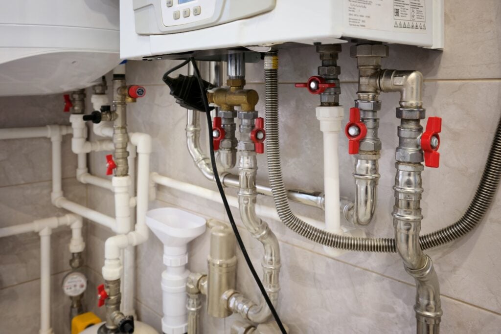 A close-up view of the complex pipe system underneath a wall-mounted boiler, featuring multiple silver metal pipes, white plastic tubes, and red shut-off valves.