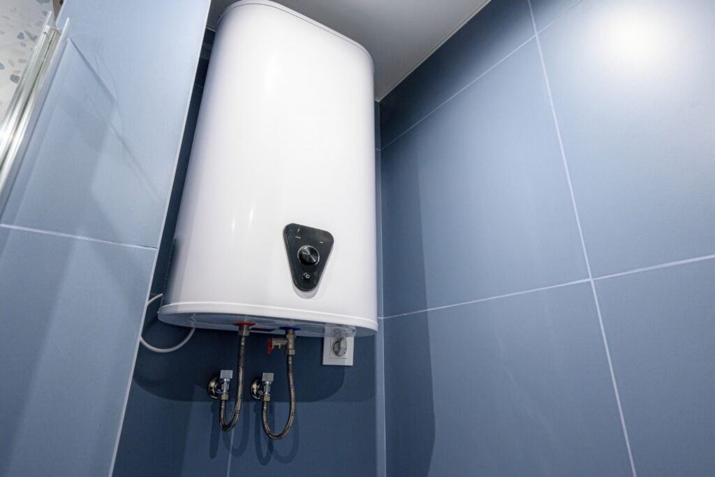 A white, wall-mounted electric water heater with a black control dial installed against blue wall tiles.